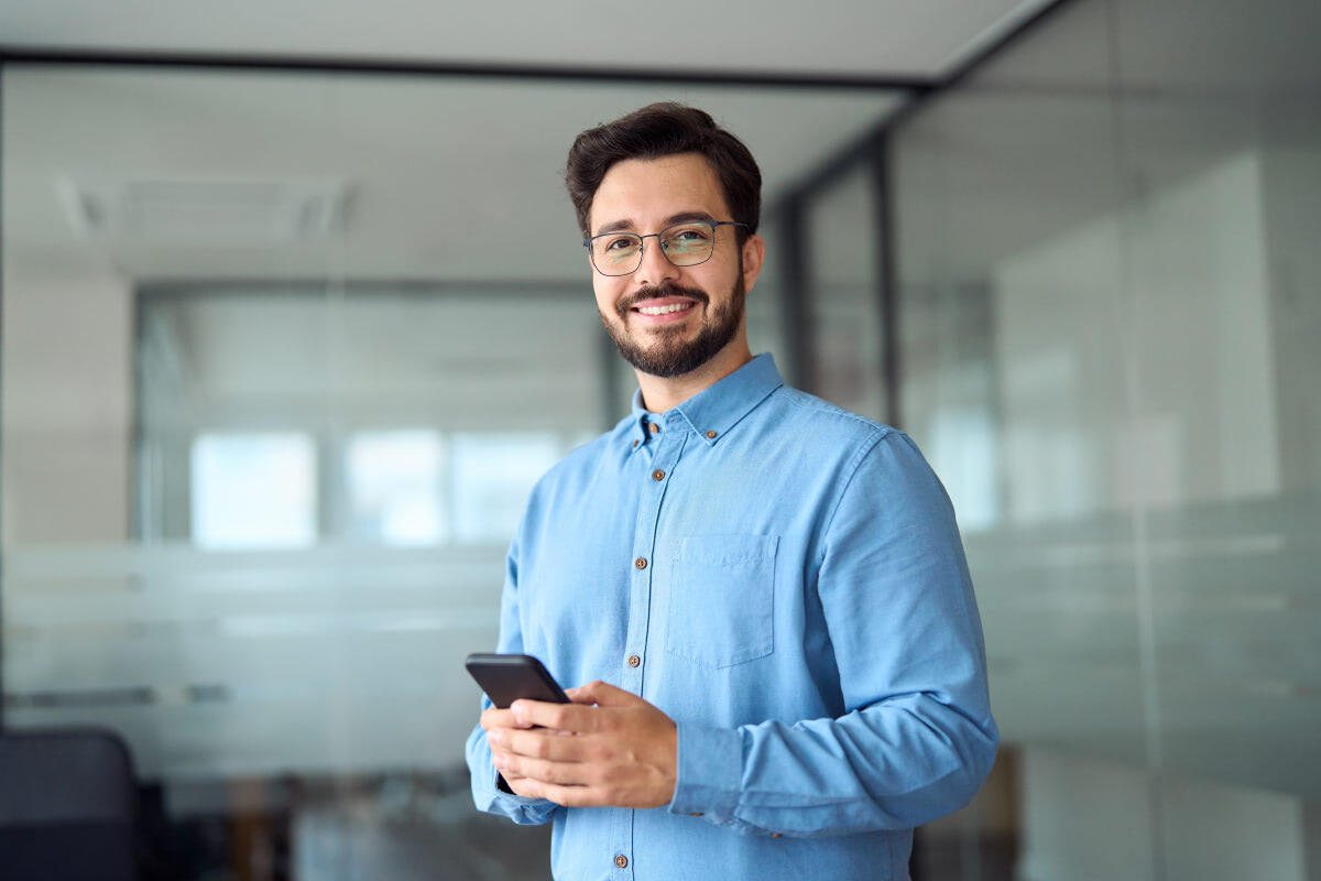 A male person with a beard and glasses holding a phone in an office